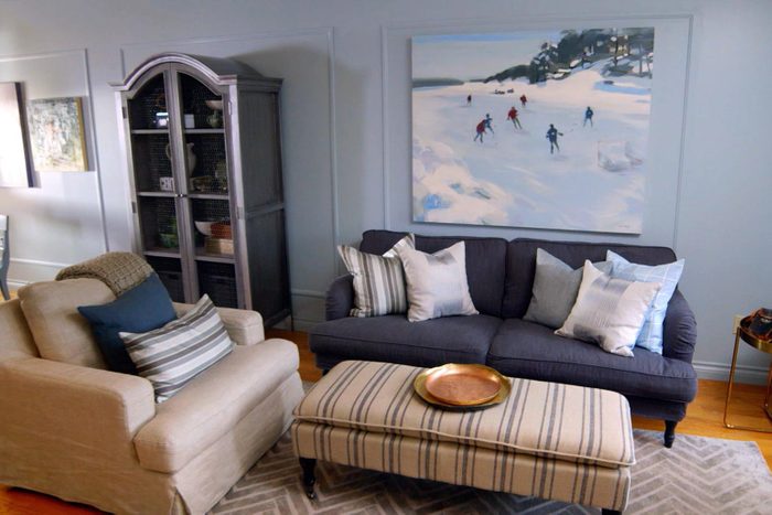 a nicely decorated living room with light blue-grey walls, a couch and arm chair with a painting of people skiing on the wall behind the couch